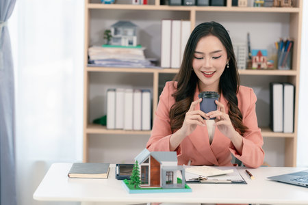 Real estate agent sitting in office workplace, concept of buying and loan in home title working, laptop computer while other hand holding model house.の写真素材
