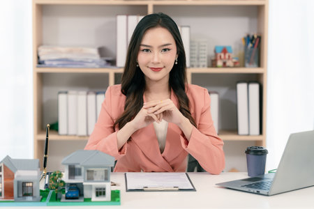 Real estate agent sitting in office workplace, concept of buying and loan in home title working, laptop computer while other hand holding model house.の写真素材