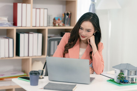 Real estate agent sitting in office workplace, concept of buying and loan in home title working, laptop computer while other hand holding model house.の写真素材