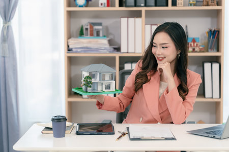 Real estate agent sitting in office workplace, concept of buying and loan in home title working, laptop computer while other hand holding model house.の写真素材
