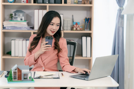 Real estate agent sitting in office workplace, concept of buying and loan in home title working, laptop computer while other hand holding model house.の写真素材