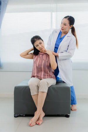 Asian female doctor examines elderly female patient with neck and shoulder pain. The doctor examines...