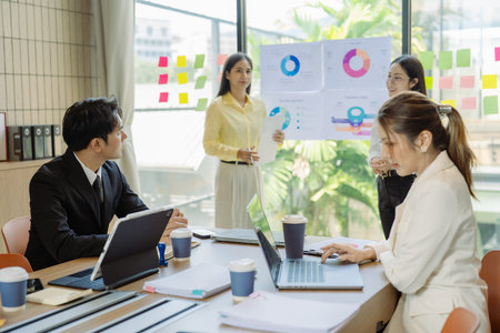 Group of Asian business people presenting data to colleagues in conference room. Business people in suits holding pens pointing at graph documents on table to present company data.の写真素材