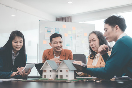 A group of young Asian businesspeople meet at an office for a residential real estate project. A group of elegant, professionally dressed individuals evaluate a small house model.の写真素材