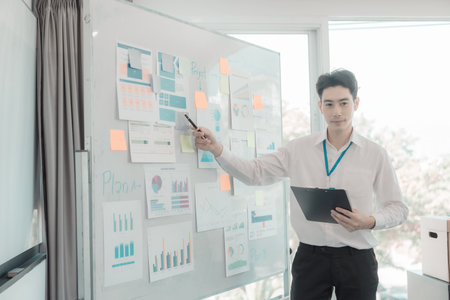 Young Asian businessman working with whiteboard in office, pointing at graph on whiteboard to present to manager in conference room, business concept.の写真素材