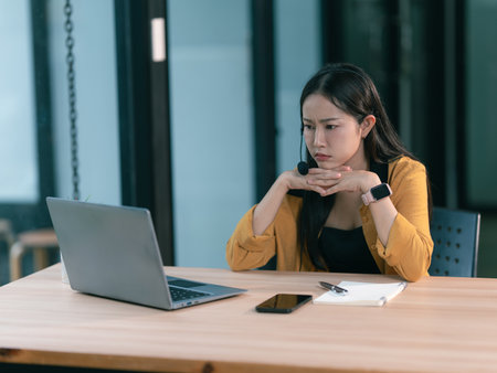 Beautiful Asian woman wearing headphones while working on laptop computer, giving advice to customers in office.の写真素材