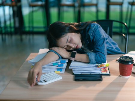 An Asian businesswoman is sleeping at her desk, looking exhausted at work with a pile of documents beside her, indicating exhaustion and hard work.の写真素材