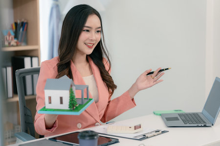 Young Asian woman sitting in office working on laptop, holding keys and house model in front for real estate concept.の写真素材