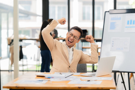 A glasses-wearing businessman is thrilled with his success while using his laptop. A happy professional is celebrating his accomplishments online. An office worker.の写真素材