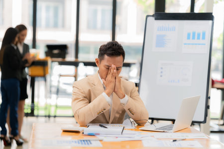 A pensive businessman wears glasses while working on his laptop at his office desk with a colleague standing behind him.の写真素材