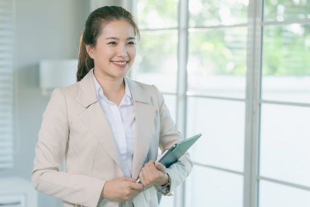 An Asian businesswoman holds a tablet while standing in an office, working online, checking emails, holding electronic devices, working and visiting websites.の写真素材