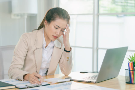 A stressed businesswoman is working on her laptop, experiencing a headache and deep thought from her office work.の写真素材