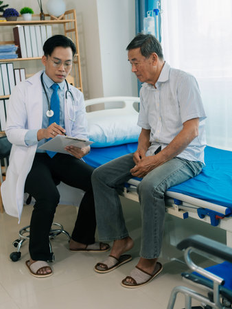 An Asian male doctor and an elderly patient are talking on a bed in a hospital room.