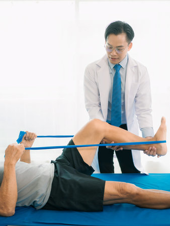 An elderly Asian man is undergoing physical therapy to stretch his legs and knees, under the care of...