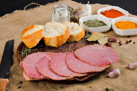 the photograph shows some pieces of ham cut into slices on top of a wooden board with some pieces of bread and some spicesの写真素材