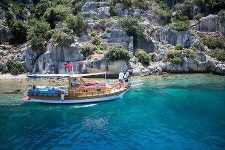 touristic boat passing by sunken city of Kekova under turkish flag close to rocksのeditorial素材