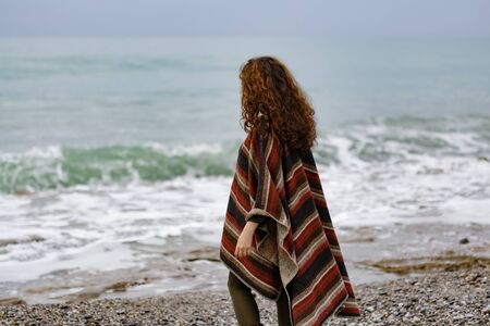 Backview portrait of happy brunette woman by seaside wearing striped red black and beige ponchoの写真素材