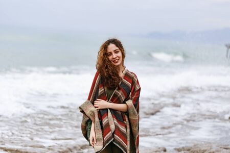 Portrait of happy brunette woman by seaside wearing striped red black and beige ponchoの写真素材
