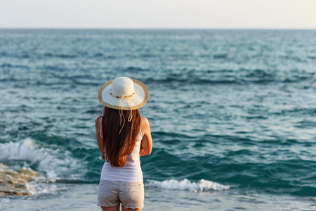 Backview portrait of young fit woman with straight long hair wearing white tshirt shorts and straw hat standing by seasideの写真素材