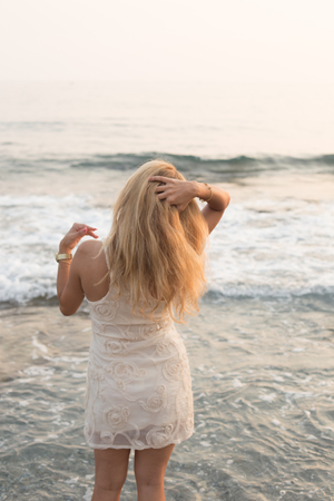 Backview portrait of a blonde woman wearing beige cocktail dress, standing by the sea and brushing her hair with handの写真素材
