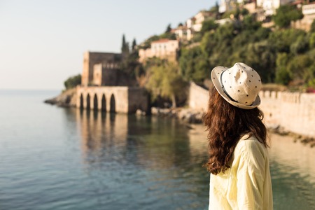 Horizontal backview portrait of a traveller woman with brown wavy hair wearing beige hat  and yellow cotton shirt enjoying the seaview and view of Mediterranean shipyard in Alanya, Turkey.の写真素材
