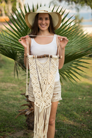 Young female with long straight hair, wearing white top and beige shorts holding a pice of macrame wall art in her hand and smiling, shot with shallow depth of fieldの写真素材