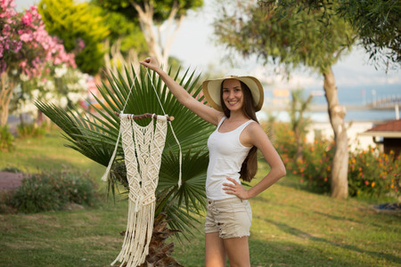 Young female with long straight hair, wearing white top and beige shorts holding a pice of macrame...