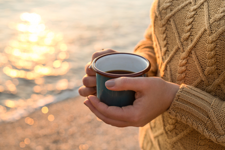 Closeup shot of female hands in cozy sweater holding dark green mug with tea at sunset light with beautiful golden bokehの写真素材