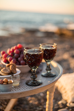 Romantic outdoor dinner with wine and cheese by seaside. Two glasses of wine, grapes, olives and dried fruits on rustic round metal table. Vertical composition.の写真素材