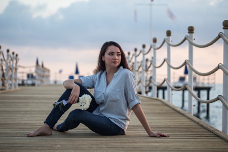 Relaxed female wearing dark blue skinny jeans and blue shirt, sitting at pier with wooden floor and holding a bouqet of white carnationsの写真素材