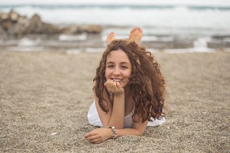 Young curly girl wearing white, lying at the beach and smiling.の写真素材