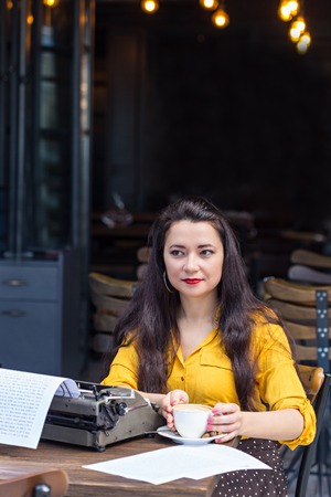Beautiful female writer with long brown hair wearing yellow shirt and polka dot brown skirt, sitting in a coffee shop with a cup of cofee in front of retro typewriterの写真素材