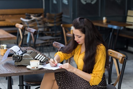 Beautiful female writer with long brown hair wearing yellow shirt and polka dot brown skirt, taking notes in a coffee shop in front of retro typewriterの写真素材