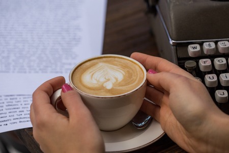 Writing concept. Closeup shot of female hands holding a cup of cappuccino in her hands with retro typewriter and papers at background.の写真素材