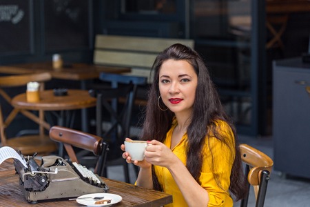 writer female with long dark hair, wearing yellow shirt and brown polka dot skirt, drinking cappuccino in a coffee shop with dark interior and with vintage typewriter on a wooden tableの写真素材