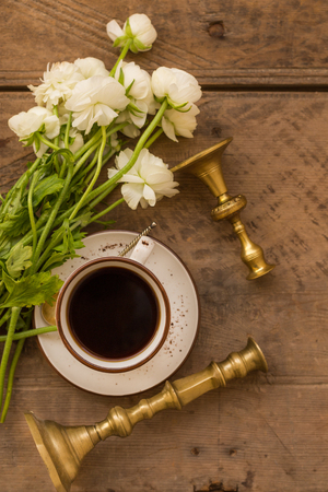 Upper view shot of a cup of coffee, white ranunculus and vintage candleholders of golden color on rustic wooden background.の写真素材