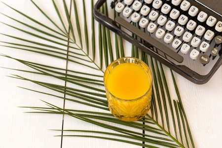 A glass of orange juice on palm tree leave with retro typewriter on white wooden tabletop.の写真素材