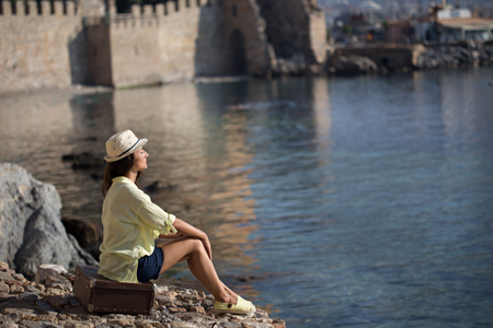 Solo traveller woman sitting on vintage suitcase by the sea and wearing dark blue shorts, yellow cotton shirt and hat. Copy space.の写真素材