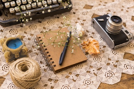 Spring or summer freelance, business or writing concept. Notepad with black pen on lace table cover with vintage film camera, seashells and candle.の写真素材