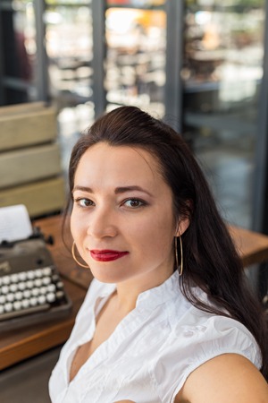 Head and shoulders portrait of self-confident writer female wearing white shirt and red lipstick, with blurred retro typewriter at far distance view. Vertical composition.の写真素材