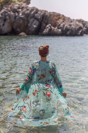 Backview portrait of female wearing colorful tunic and head wrap, entering pristine water of the secluded bayの写真素材
