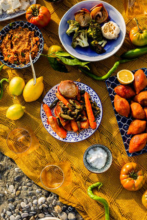 Topview image of a colorful beach picnic with grilled carrots, brokkoli, onions and garlic, bulgur, yogurt, staffed meatballs served in a blue and white ceramic tableware on a yellow blanketの写真素材