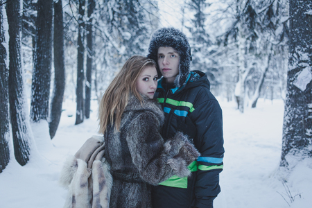 beautiful girl with flowing hair in a gray coat and a guy in a black jacket and hat against the background of the winter forestの写真素材