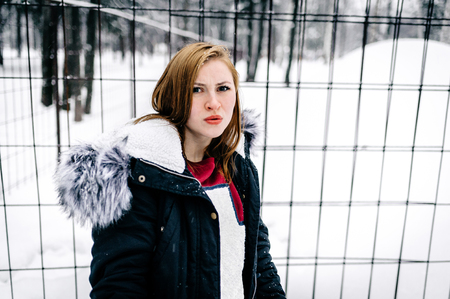 the beautiful girl with displeased face beside the fence of metal bars amid winter forestの写真素材