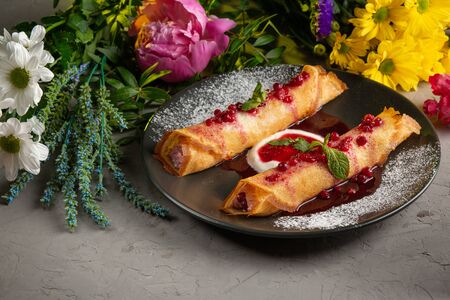 pancakes stuffed with cottage cheese with sour cream and lingonberry sauce in a black plate on a gray background and flowers in the backgroundの写真素材