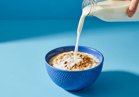 A close-up shot captures white milk streaming from a clear bottle, being poured over a textured blue bowl filled with creamy oatmeal and sprinkled with granola.の素材