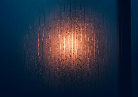 Close-up view of water droplets clinging to a glass surface, illuminated by a warm, soft light creating an abstract and moody atmosphere.の素材