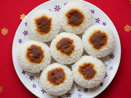 A white plate with purple flowers holds seven round rice balls with brown sauce centers on a red tablecloth with yellow and orange flowersの素材