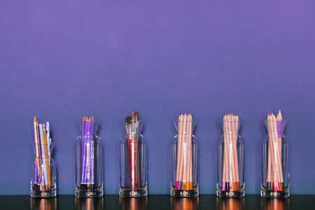 Artistic tools. Clear glass cups lined with colored pencils and paint brushes on dark wood and purple backgroundの写真素材