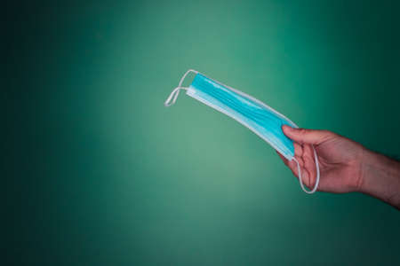 hand of a young adult man holding a light blue disposable mask with light blue folds with white strips on a sea green background alluding to the theme of the virusの写真素材